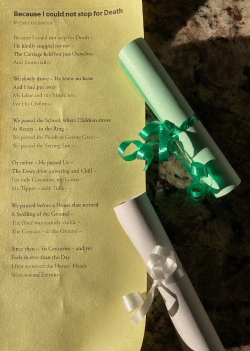 An unrolled paper scroll with Emily Dickinson's "Because I could not stop for Death" printed on it with two more scrolls tied with ribbons on the right side of the image