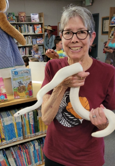 A woman with glasses and gray hair holds a white snake in front of library shelves full of books