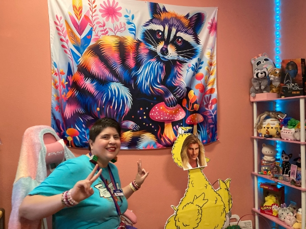 A woman in a teal shirt in front of a colorful raccacooni wall hanging and a set of shelves packed with fun objects