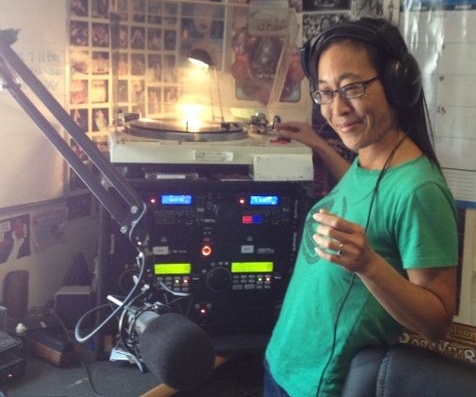 DJ FlutterByCam wearing a green shirt and headphones in a community radio studio while playing a vinyl record