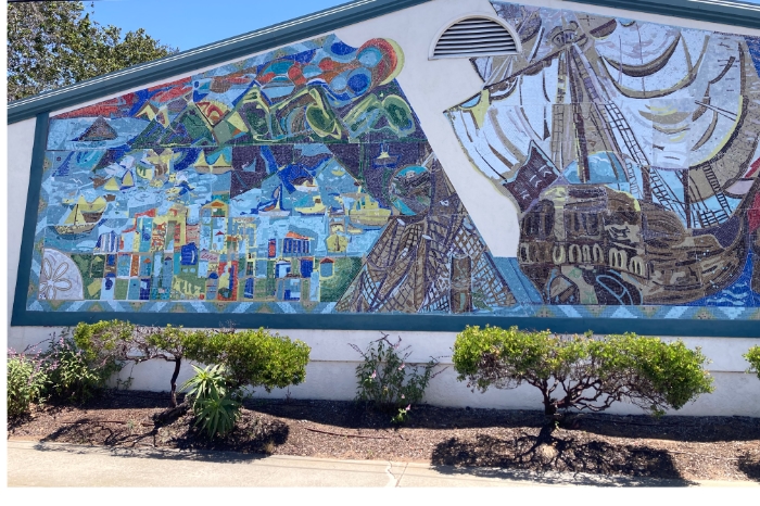 A colorful mosaic mural on the exterior wall of a building featuring a sailing ship, Morro Rock, the fishing village of Morro Bay, and a stylized Halley's Comet